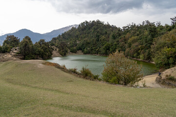 A tranquil lake surrounded by lush green hills and dense forests under a cloudy sky. A walking path and grassy landscape enhance the natural beauty of this serene and untouched trek in Uttarakhand.