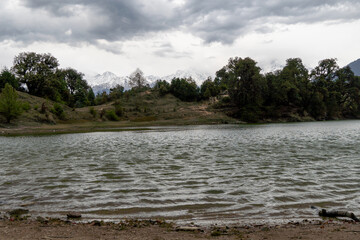 A serene lake with gentle ripples reflects the cloudy sky, surrounded by dense green trees and distant snow-capped mountains. Peaceful overcast atmosphere at Deoria tal lake trek in Ukhimath, India.
