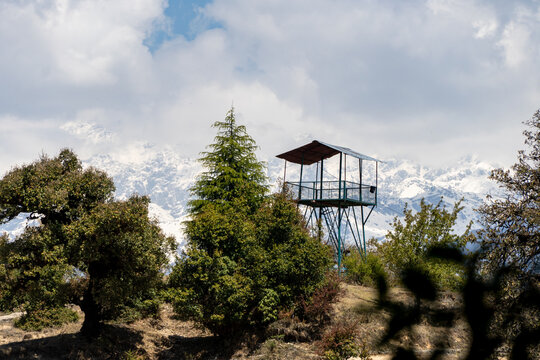 A watchtower stands among green and tall cedar trees and snow-capped mountains visible in the background under a cloudy sky. Mesmerizing view while trekking to Deoria tal lake in Uttarakhand, India.