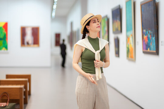 Mid shot of a young Caucasian woman in elegant jacket, straw hat and trousers contemplating in the art gallery. Painting in background. Concept of cultural education and Museum Day