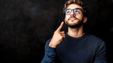Obraz premium A young man with glasses and a beard displays a thoughtful expression while pointing upwards in a dim background, evoking emotions of inspiration and contemplation.