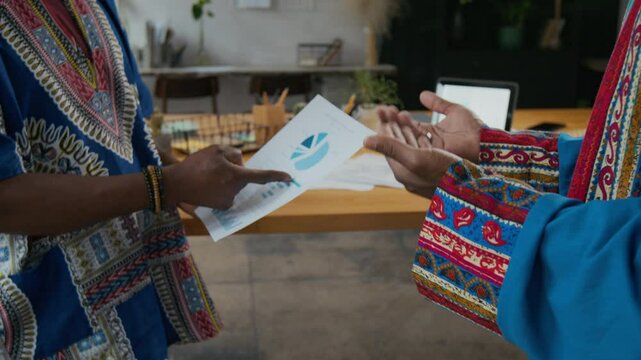 Close-up shot of unrecognizable Black men in traditional African outfits discussing business documents and shaking hands in agreement in modern office