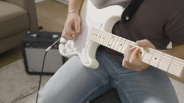 Slow motion of a Young man playing a riff on a white Stratocaster style electric guitar with a small amplifier in the background.