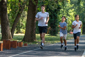 Fototapeta premium Trainer runs alongside two boys in a sunny park, focusing on improving their endurance and physical fitness. The boys are energized, learning proper running techniques while enjoying outdoor exercise