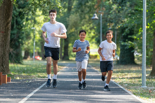 Trainer runs alongside two boys in a sunny park, focusing on improving their endurance and physical fitness. The boys are energized, learning proper running techniques while enjoying outdoor exercise - Powered by Adobe