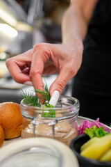 A person's hand adds fresh dill to a jar of spread in a kitchen setting. The background features various ingredients, including rolls and pickled vegetables, highlighting a culinary preparation.