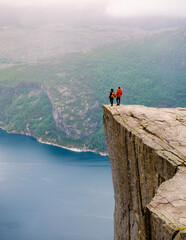 Two individuals stand on the edge of Preikestolen, a dramatic cliff in Norway, gazing out at the...