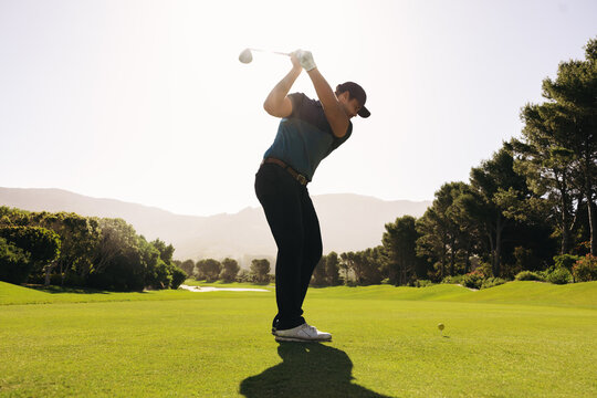 Man golfer swinging club at golf ball on fairway shot under sunny sky surrounded by lush greenery