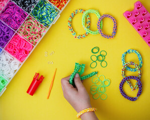 Child's hand holding a work in progress loom bracelet, other colorful elastic bracelets and bracelet making supplies on yellow background. Toy developing children's imagination. Flat lay, from above.