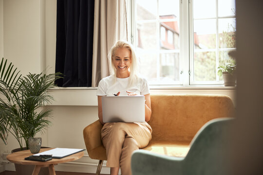Psychologist speaking with a client online. She sits on a yellow sofa and looks into the camera on her laptop smiling and engaged.