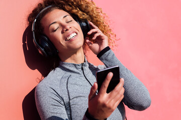 cheerful sportswoman with mobile phone listening to music from headphones against pink wall, concept of technology of communication and urban lifestyle, copy space for text