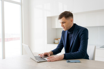 Man in a suit is typing on a laptop. He is sitting at a table with a cell phone and a cell phone charger