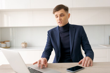 Man in a suit is sitting at a table with a laptop and a cell phone. He is looking at the laptop screen