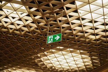 A green emergency exit sign hangs from a complex geometric ceiling. The intricate design and...