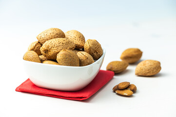 Almonds in white bowl, next to shelled almonds, on white wooden surface.