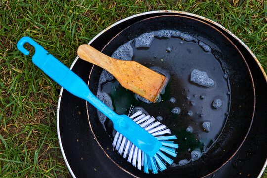 Rinsing kitchenware outdoors. Dirty frying pan with soap foam, a wooden spatula, and a blue brush on green grass. Minimalistic camping lifestyle,  washing and clining. 
