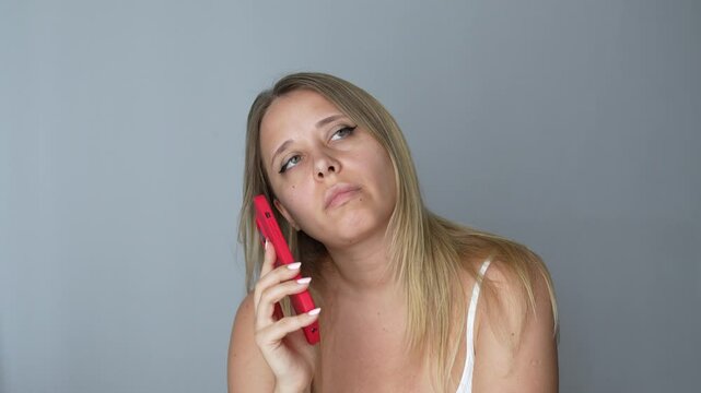 Young bored blonde woman holding the mobile phone in red case listening to long speeches or voice messages rolling her eyes on a dark grey color background. Long wait on the line