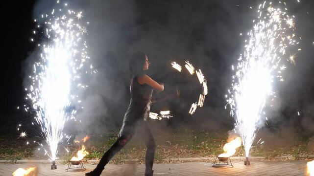 The silhouette of a circus performer with burning torches in her hands on a black background, the performance of artists with fire and fireworks fountains. Artists staged a night show waving fireballs