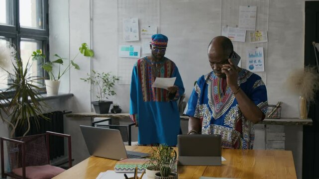 Young Black man in vibrant African outfit interrupting his senior colleague from phone call to examine documents in modern open space office