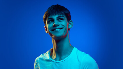 Portrait of handsome young man,s student in white t-shirt, looking upward and smiling against blue studio background in neon light. Happiness and dreams. Concept of youth, emotions, lifestyle, fashion