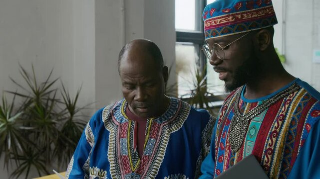 Tilt-up shot of two Black men in traditional African blue dashikis pensively analyzing business documents in modern office