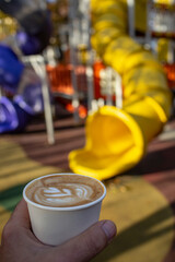 Male handholding a disposable paper cup with a latte art flat white with colorful defocused playground in the background