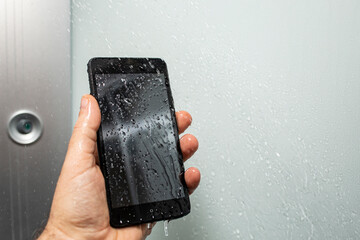 Male hand holding a black smartphone under shower with water drops and motion blur, soft focus...