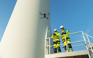 Two wind turbine or windmill workers or engineer stand on base of the pole and one control drone with discuss together in workplace field.