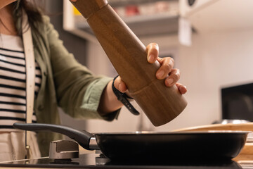 Female hand using a wooden pepper mill over a frying pan in modern kitchen
