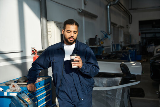 Handsome mechanic takes a moment to relax with a warm drink amidst busy workstations.