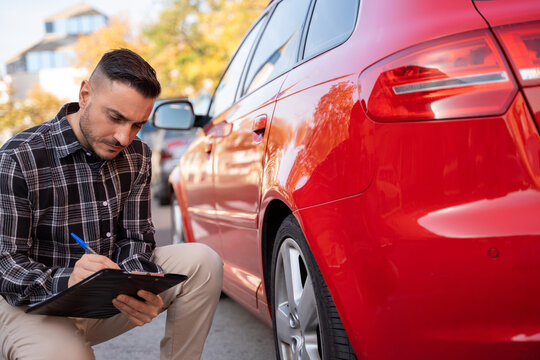 Insurance agent crouching near a damaged red car and writing on a clipboard, assessing the damage for the insurance claim