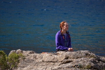 Fototapeta premium A young girl sits on a rock in the middle of a blue mountain lake
