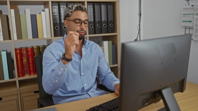 Young bearded man in an office room stretching and taking off his glasses at a desk with a computer, looking exhausted with bookshelves in the background