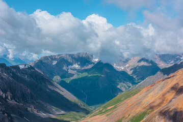 Fototapeta premium Scenic aerial view to beautiful colorful alpine valley among multicolor mountain ranges with snow in sunlight in low clouds under blue sky. Vivid landscape with shadows of clouds on high mountains.