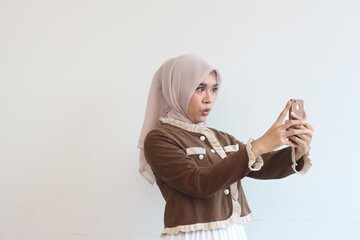 A Young Woman in a Brown Cardigan Takes a Selfie with Her Smartphone Against a White Background