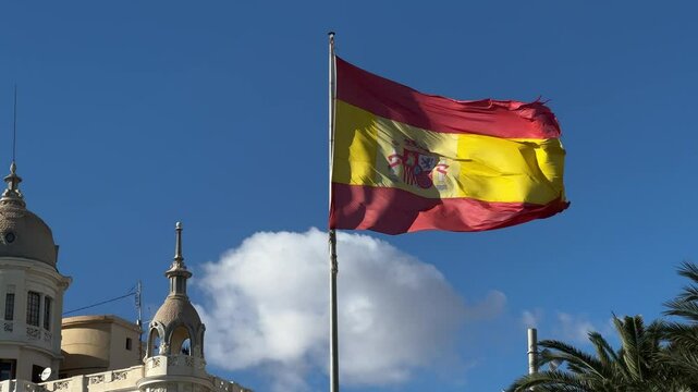 Spanish flag waving against blue sky with architectural domes and palm trees in Alicante, Valencia, Spain