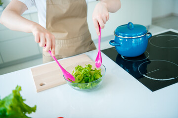 Young woman in casual style apron preparing fresh salad in a modern kitchen with pink utensils