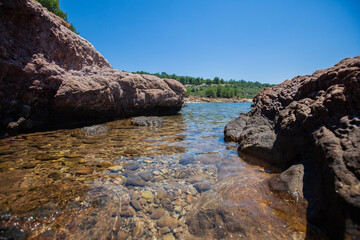 tranquil rocky shoreline featuring smooth pebbles beneath crystal clear water. Framed by rugged...