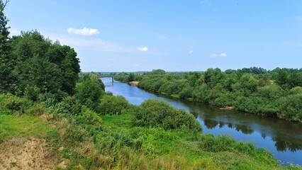 Fototapeta premium A bridge on concrete piers rises above the river near a sandy beach. Shrubs and deciduous trees grow along the grassy banks. Sunny summer weather