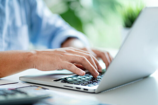 A man is typing on a laptop computer. He is wearing a blue shirt. The laptop is open and the keyboard is visible