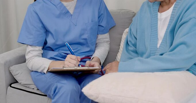 Closeup of Caucasian female doctor in blue scrubs taking notes during consultation with Asian elderly patient sitting on couch concept detailed medical care documentation trust communication