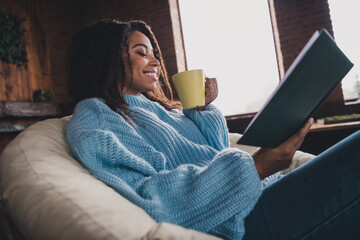 Young woman enjoying a cozy day indoors while sipping coffee and reading a book in a blue knitted...