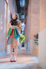 Fototapeta premium Stylish young woman holding colorful shopping bags while admiring the architecture of a charming brick alley, enjoying a pleasant day of retail therapy