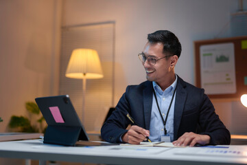 Happy businessman taking notes while attending a video conference call on a tablet in his office at night