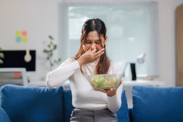 Young businesswoman is pinching her nose while holding a bowl of spoiled salad, showing a disgusted...
