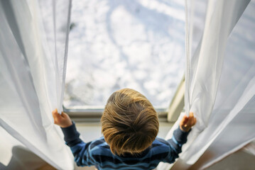 Overhead view of a blonde Boy opening net curtains and looking out of a window at winter snow