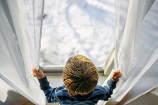 Overhead view of a blonde Boy opening net curtains and looking out of a window at winter snow