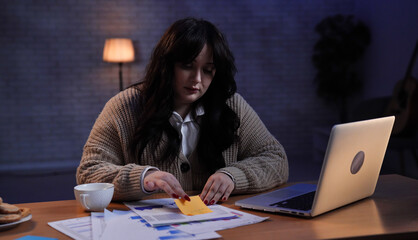 Young business woman freelancer sitting at desk working on remote in home office late, reading documents, sticking colorful memo paper on page.