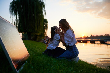 Mother and her daughter share tender moment by lakeside at sunset, with solar panel nearby. Harmony between renewable energy and natural beauty, promoting sustainable future.