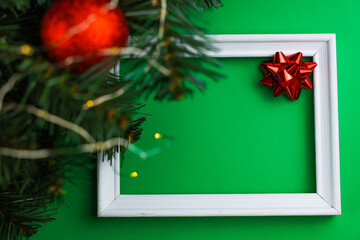A white wooden frame lies in the center of the image, with a red bow in the right corner and on the left of the image you can see the branches of a Christmas tree with a red shiny ball and a yellow